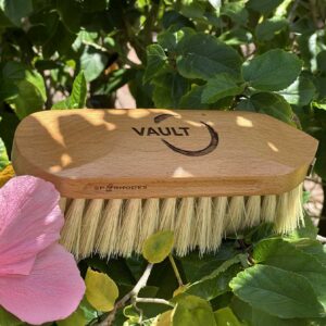 Engraved wooden dandy brush with custom engraved logo, featuring natural bristles and SP Rhodes branding, displayed among green leaves in outdoor equestrian setting